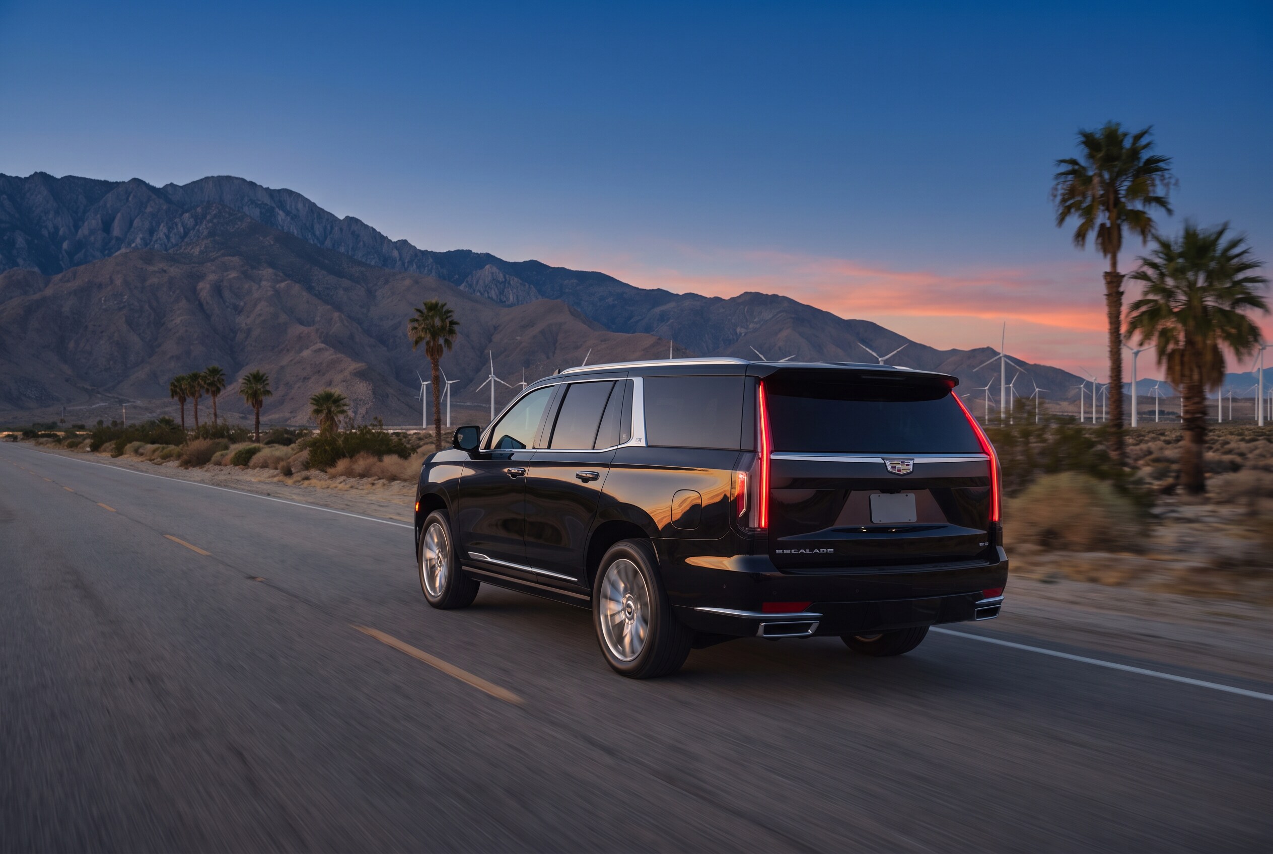 Black Cadillac Escalade on a desert highway with wind turbines and mountains at dusk